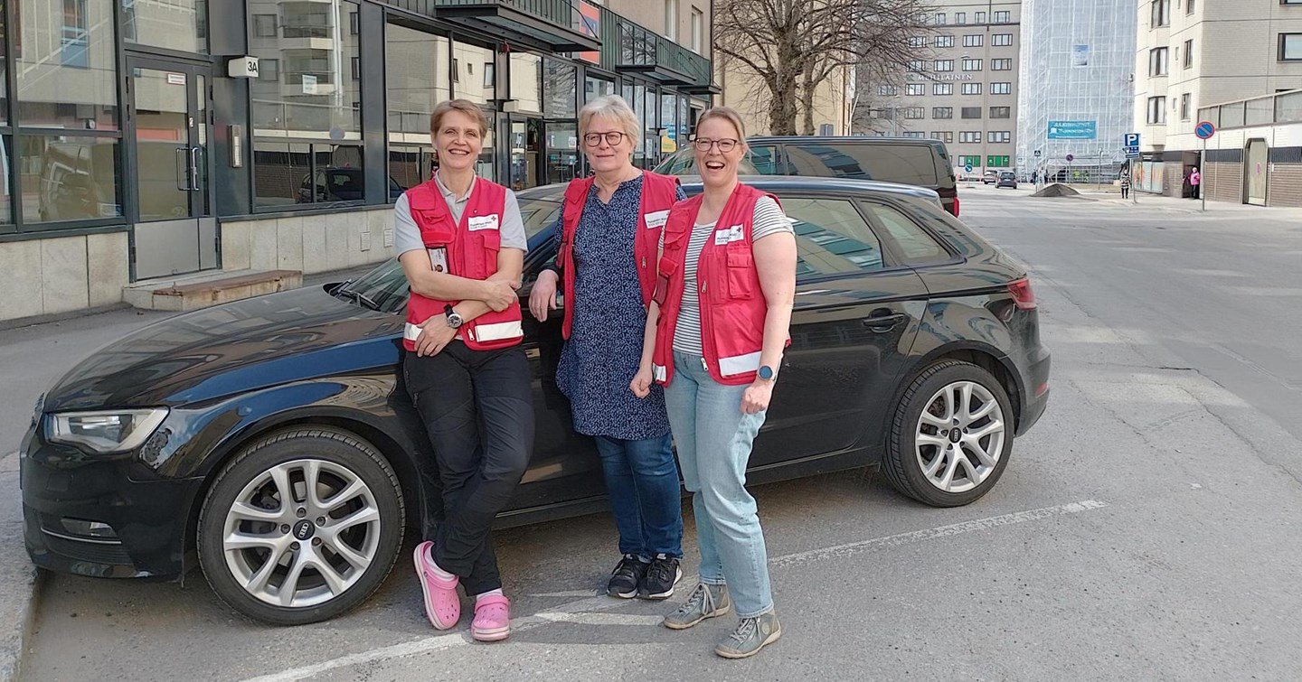 Three smiling people in Red Cross clothing standing in front of a car.