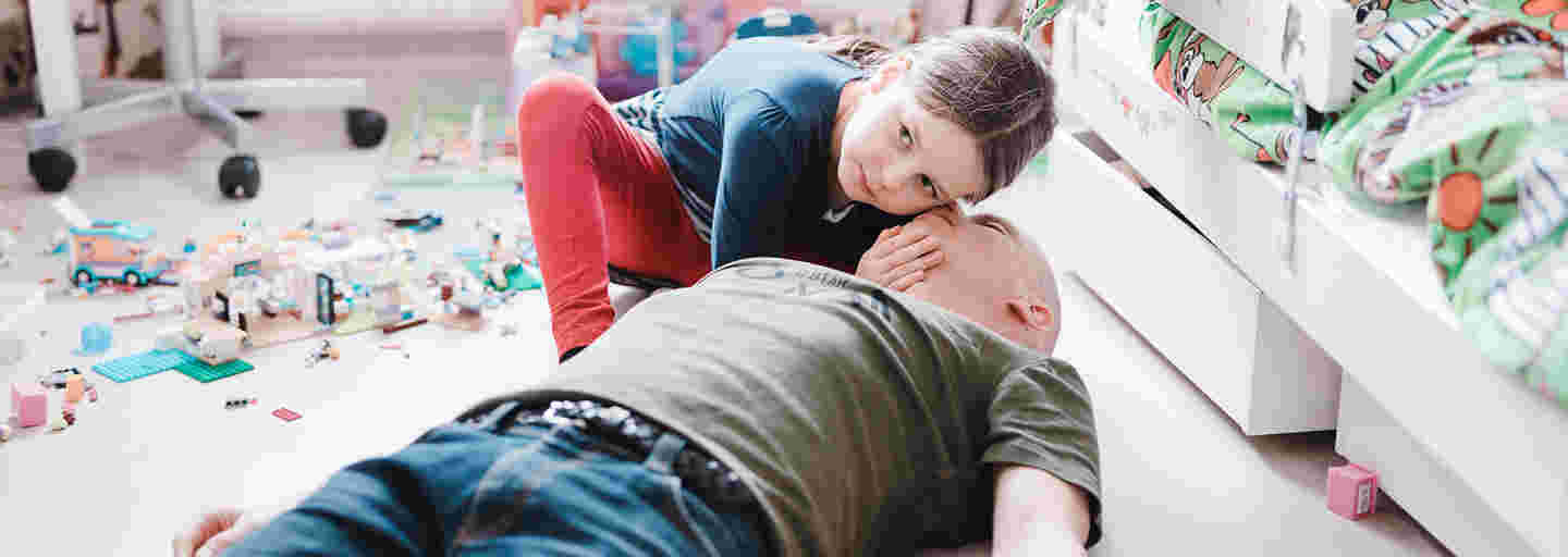 A man is lying on his back on the children’s room floor and a girl is checking his breathing with her ear to his mouth.