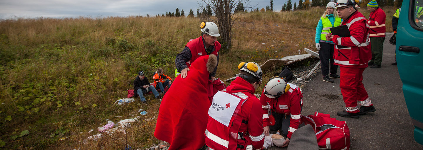 Two Red Cross ski slope volunteers wrapping a blanket around someone lying on the snow at the side of a ski slope.