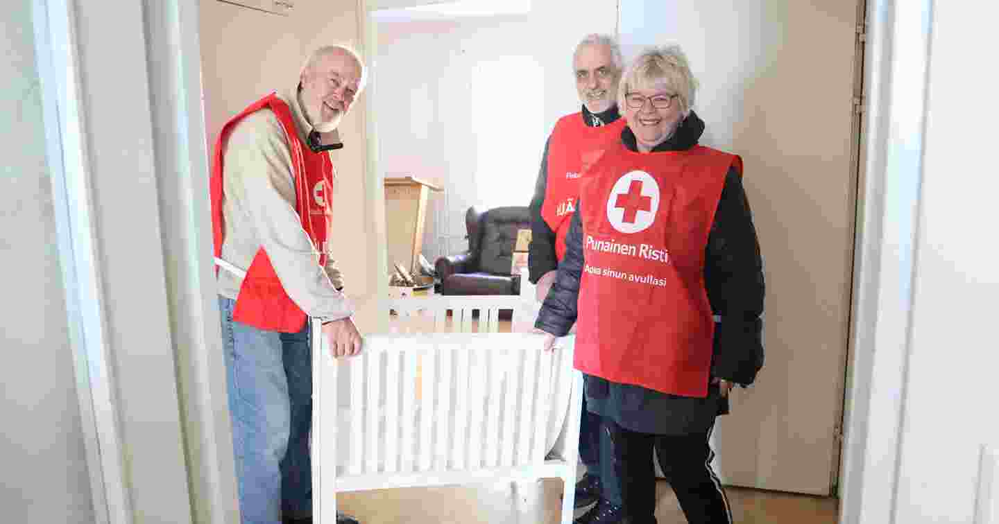 Three smiling people wearing Red Cross volunteer vests stand next to an infant bed in a dwelling that they have furnished for refugees.
