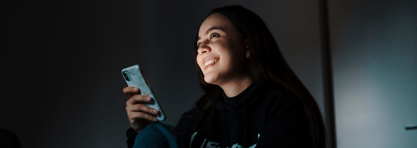 A smiling young volunteer is sitting with a phone in hand. The image is dark, but light falls on their face.