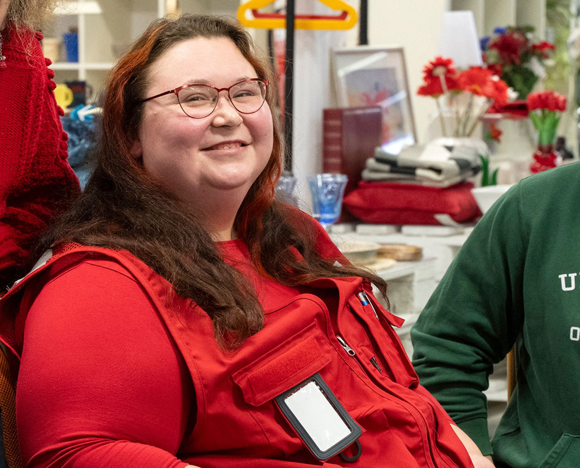 Krista Ananin, smiling while seated indoors, wearing a red volunteer vest and glasses, with shelves and red flowers in the background.