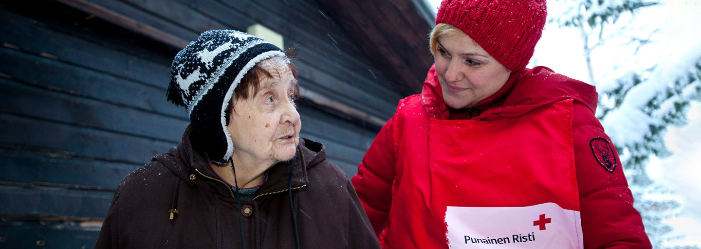 Two women standing side by side in a wintry landscape. The younger woman, wearing a Red Cross volunteer vest, is looking gently at the older woman.