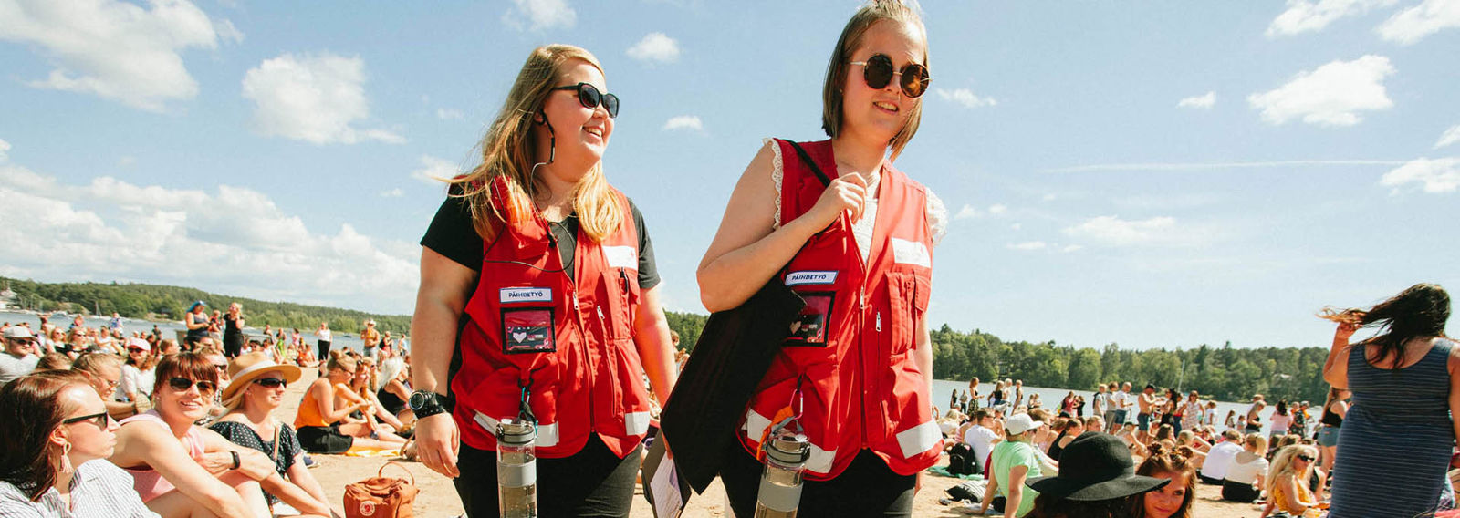 Two youths walking among seated people, wearing red vests. A summery landscape in the background.