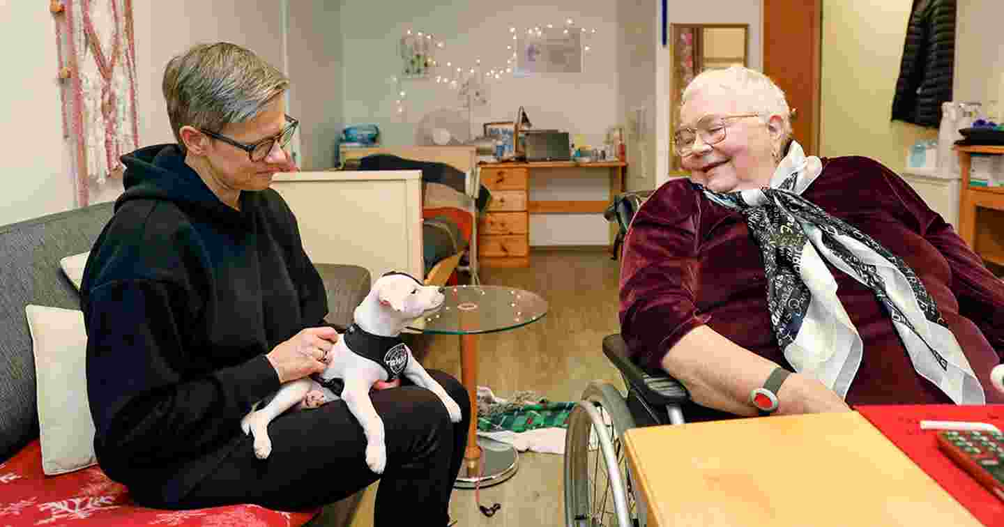 A volunteer and an older person sit around a table and smile. The volunteer has a little dog on their lap.