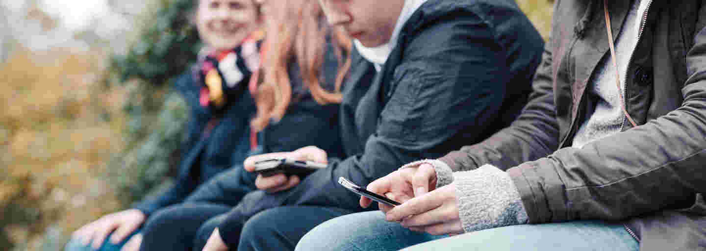 Four young people sitting side by side outside with smartphones in their hands.