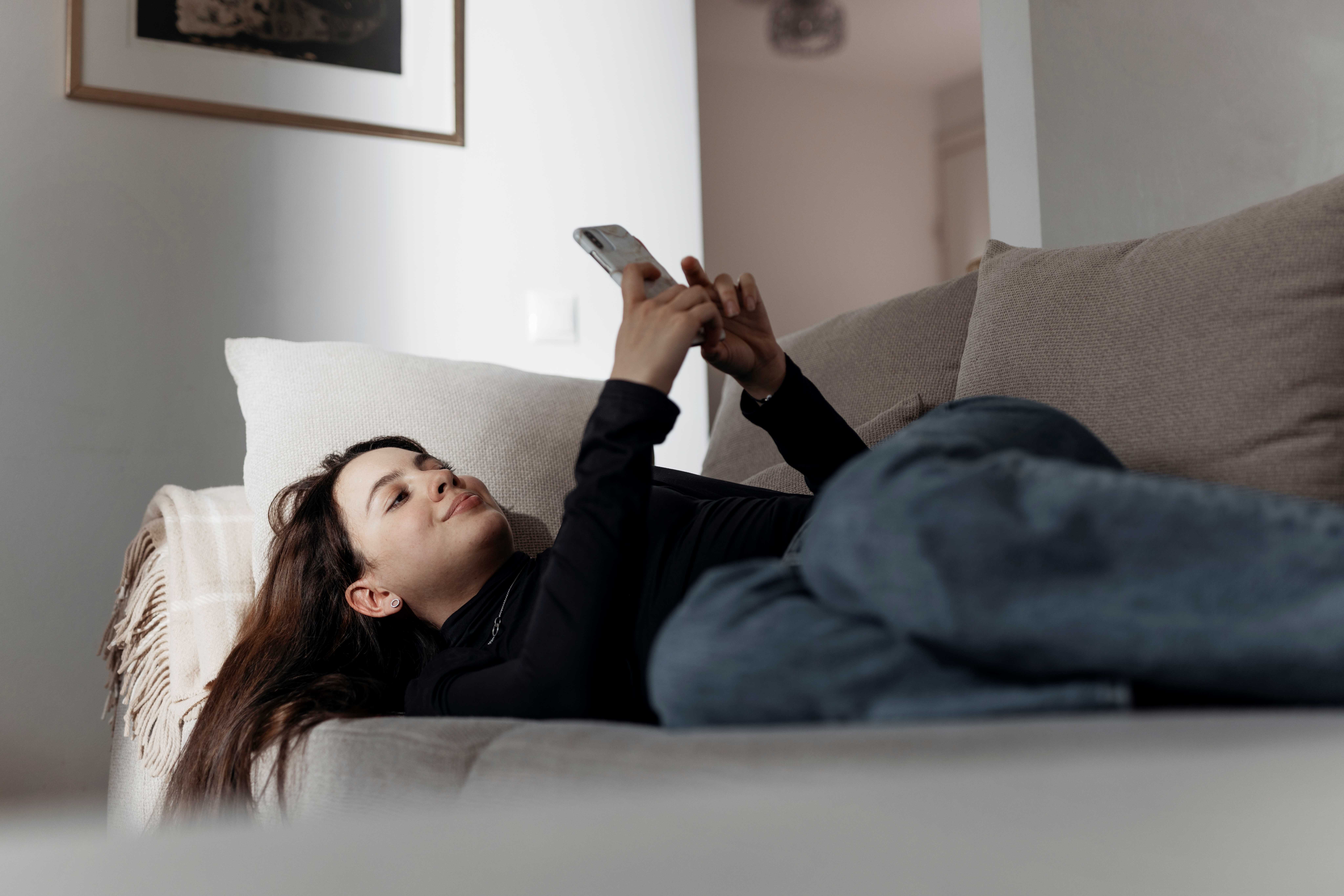 A young person is lying on the couch and scrolling through their phone.