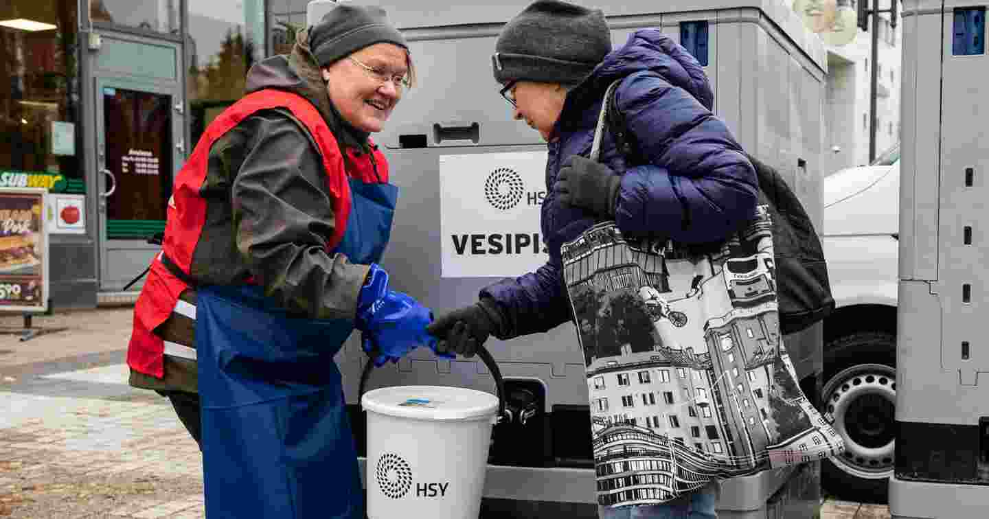 A smiling woman wearing a Red Cross volunteer vest handing a lid-covered bucket to a woman whose water supply is stopped. The Finnish Red Cross performs water distribution in domestic crisis situations.