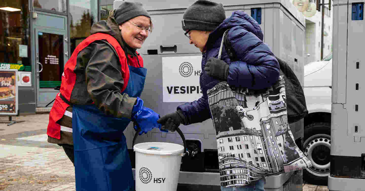 A smiling woman wearing a Red Cross volunteer vest handing a lid-covered bucket to a woman whose water supply is stopped. The Finnish Red Cross performs water distribution in domestic crisis situations.