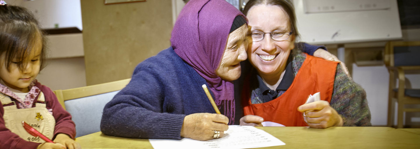 A woman wearing a Red Cross volunteer vest guiding an elderly immigrant woman and child. The elderly woman is hugging the volunteer.
