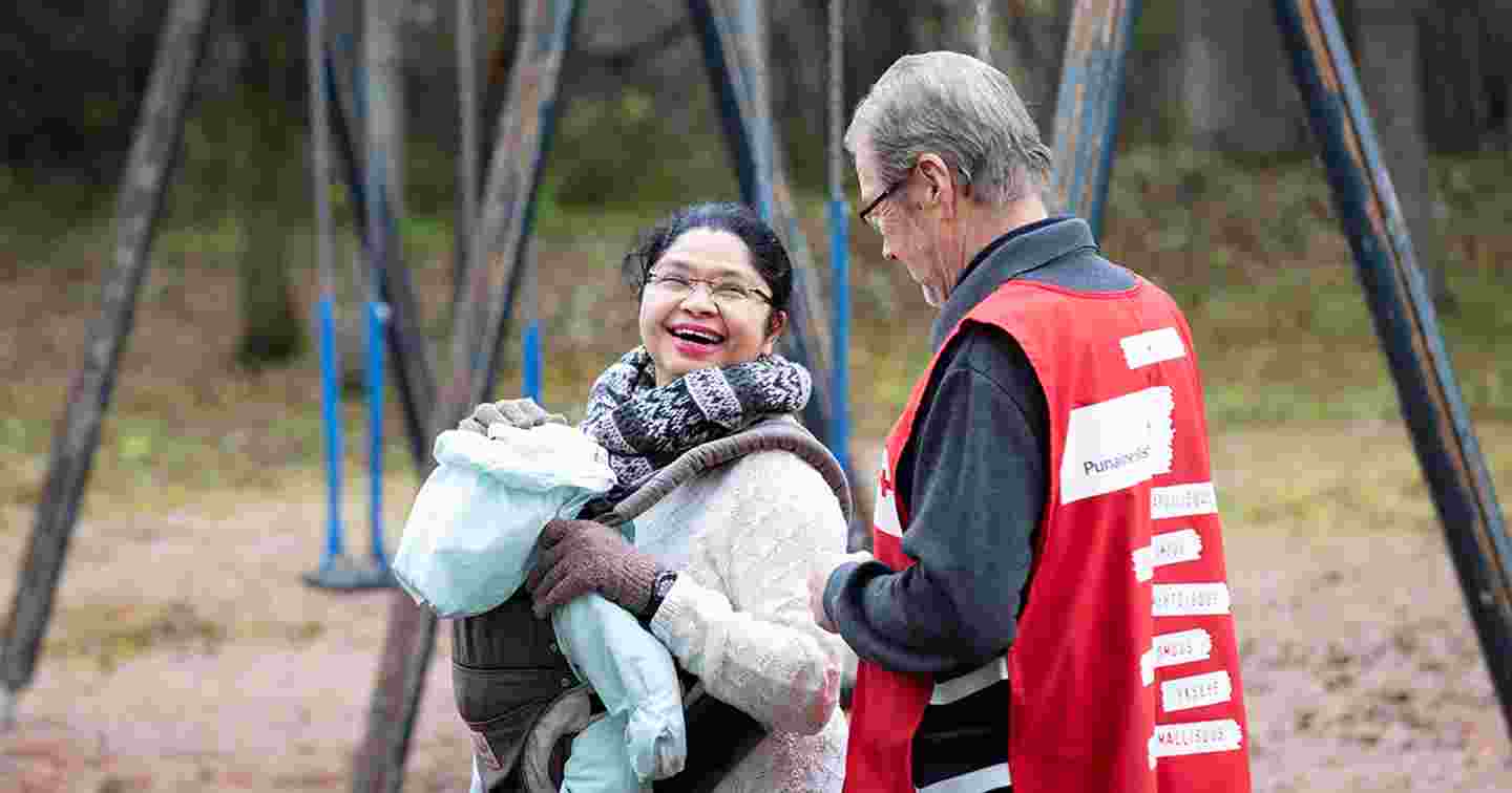A woman wearing a Red Cross volunteer vest guiding an elderly immigrant woman and child. The elderly woman is hugging the volunteer.