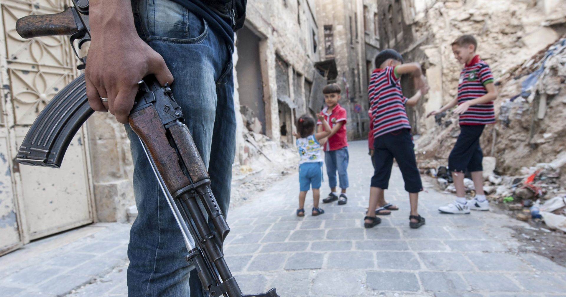 A person with a rifle in hand, children playing in the background. 