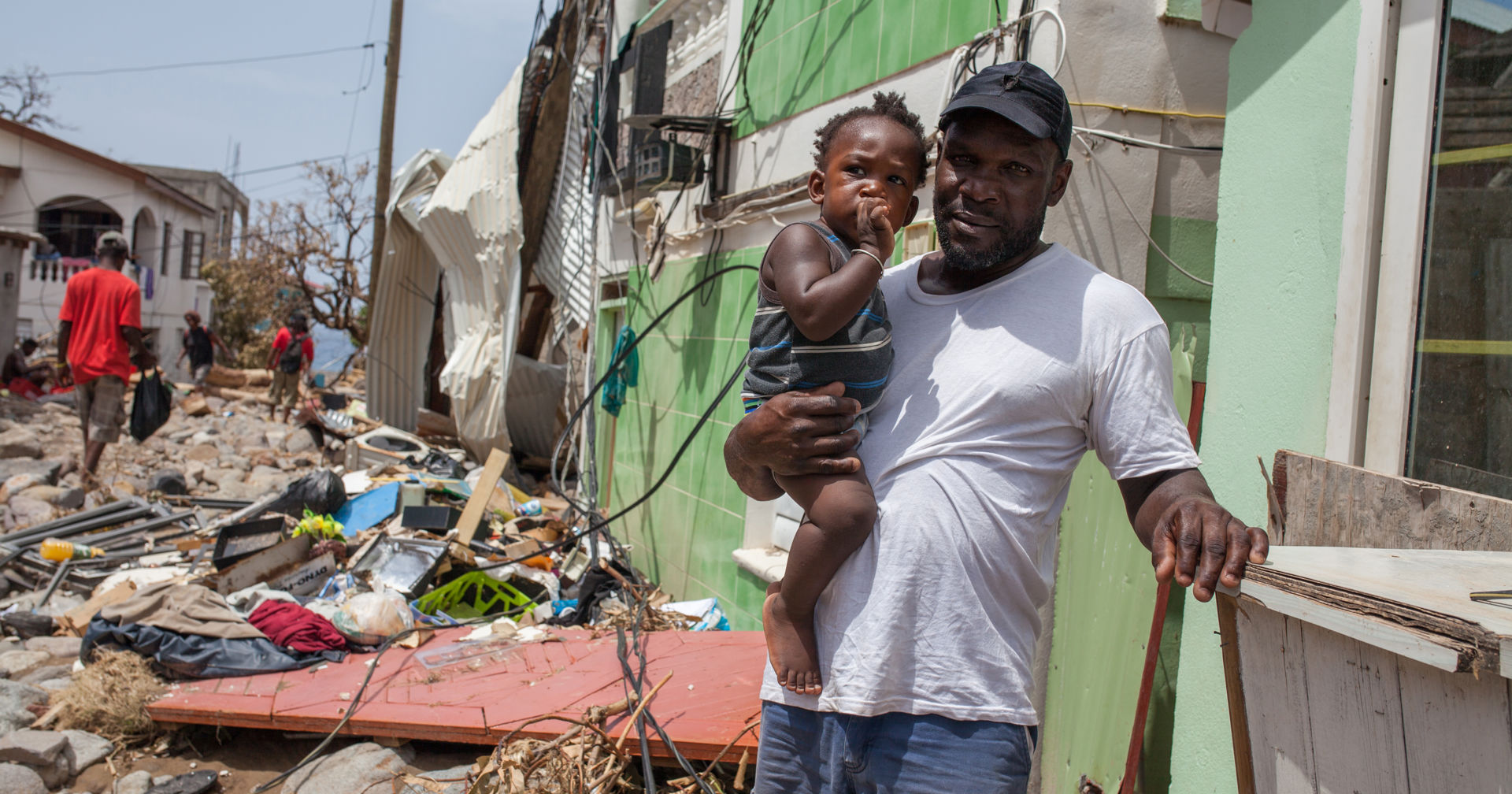 A person is holding a small child in his arms in front of a building located in the midst of storm damage.