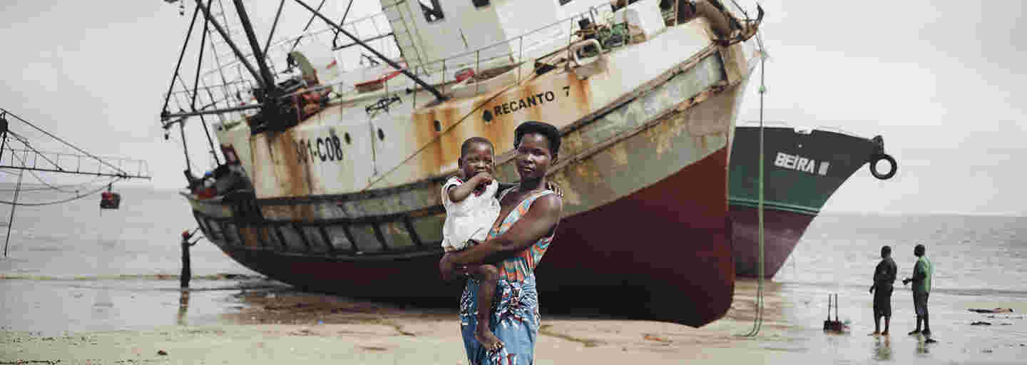 A mother standing and holding a small child in her lap on a beach. In the background, there are two ships driven ashore in a storm.