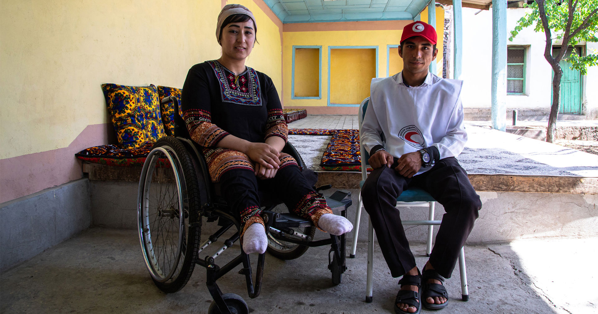 A person in a wheelchair and a person wearing Red Crescent insignia are sitting outdoors on a terrace.