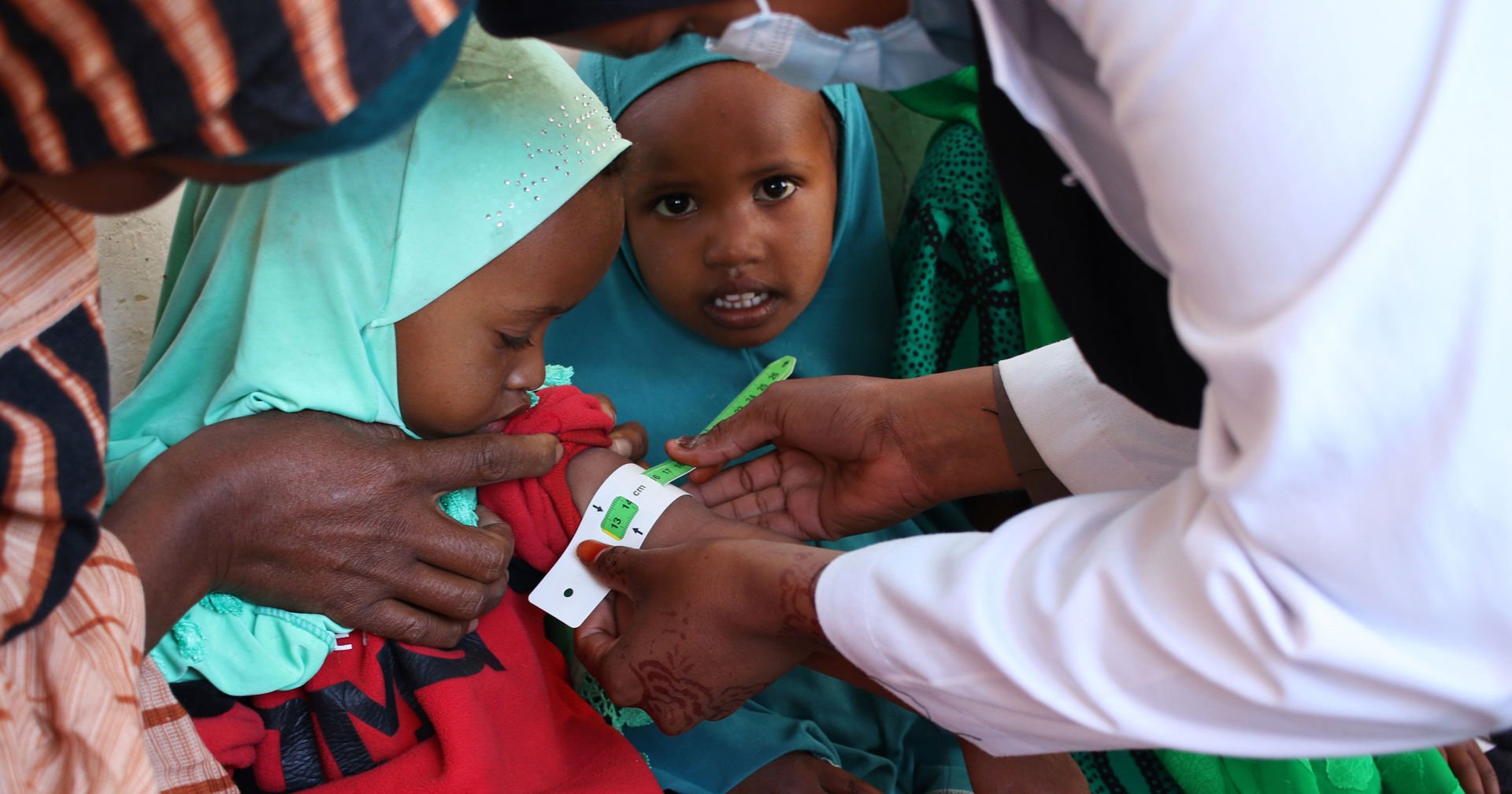 A healthcare worker measuring a child's upper arm circumference with a coloured measuring tape for nutrition screening. The child is sitting on an adult's lap, with colourful clothes in the background.