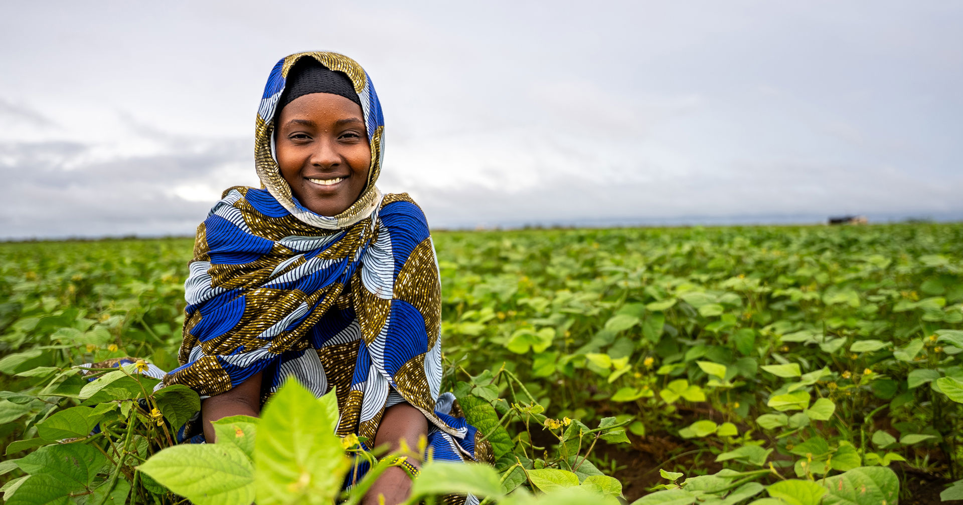 A person wearing a headscarf looks at the camera and smiles in a green field.