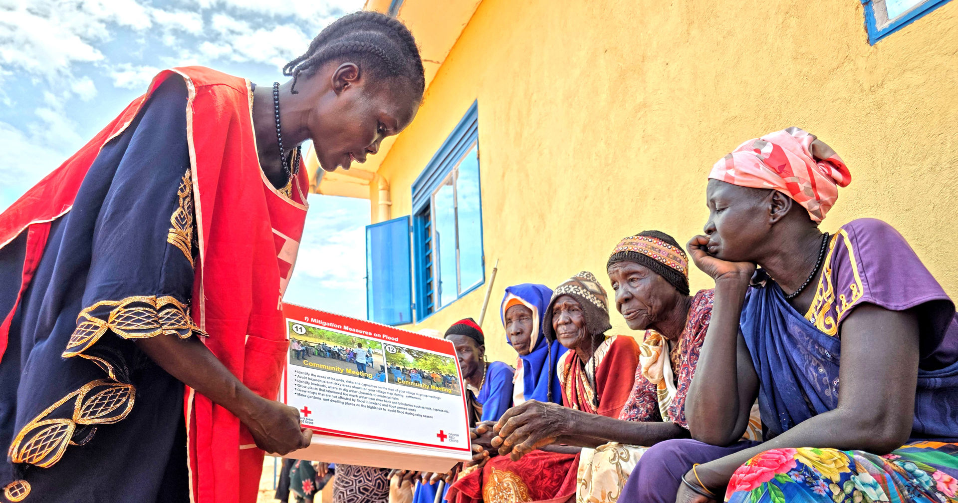 People sitting next to a yellow building, and a person dressed in a Red Cross vest shows them photos and explains what is happening in them.)