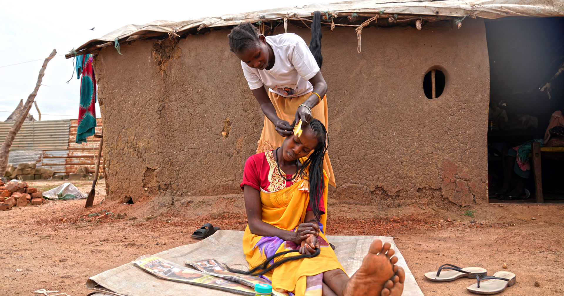  A person braiding the hair of a person sitting on top of a mat. A building made of clay is in the background.