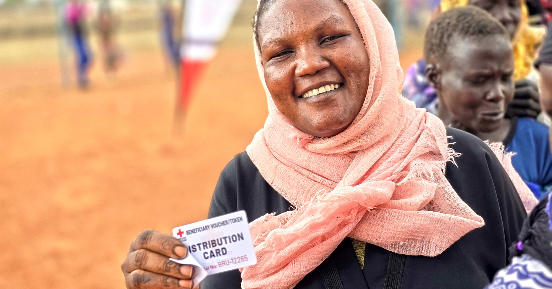 A person standing outside in a line, holding a cash assistance distribution card with the Red Cross logo.