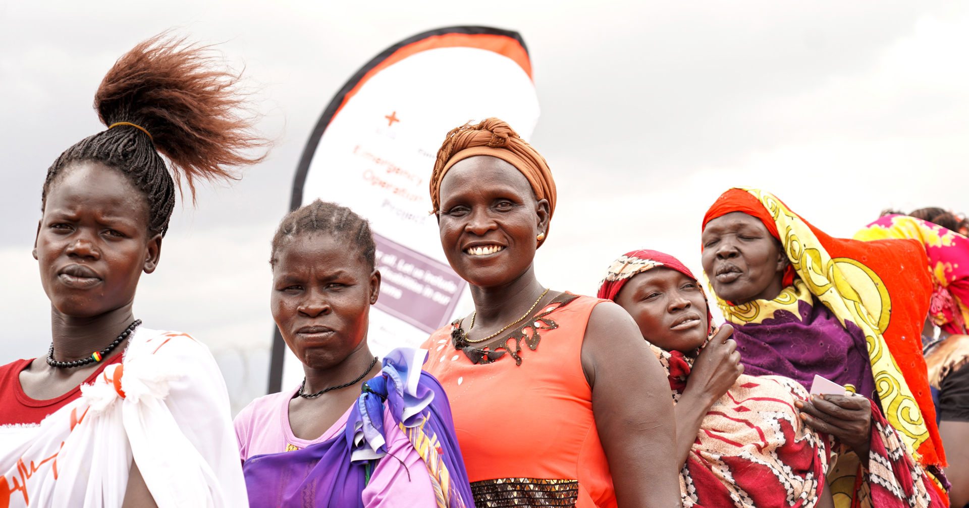 People standing in a line outside in windy weather. In the background, there is a banner with the Red Cross logo.)