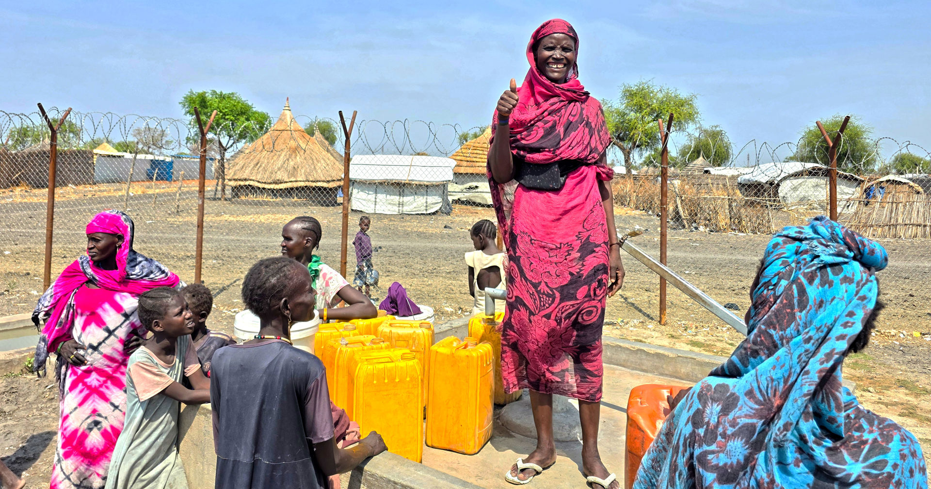 People have gathered around a water point operated with a manual pump, holding yellow plastic canisters. One person is standing on top of the well and showing thumbs up.)