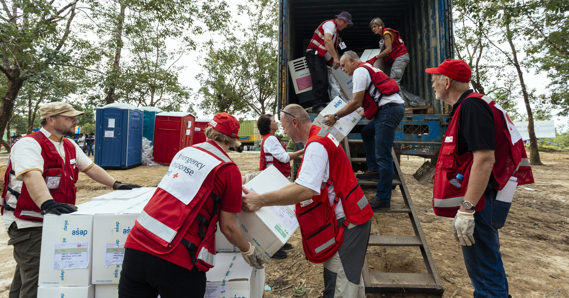 A group of Red Cross aid workers unloading boxes of relief supplies from a shipping container and passing them along on the ground.