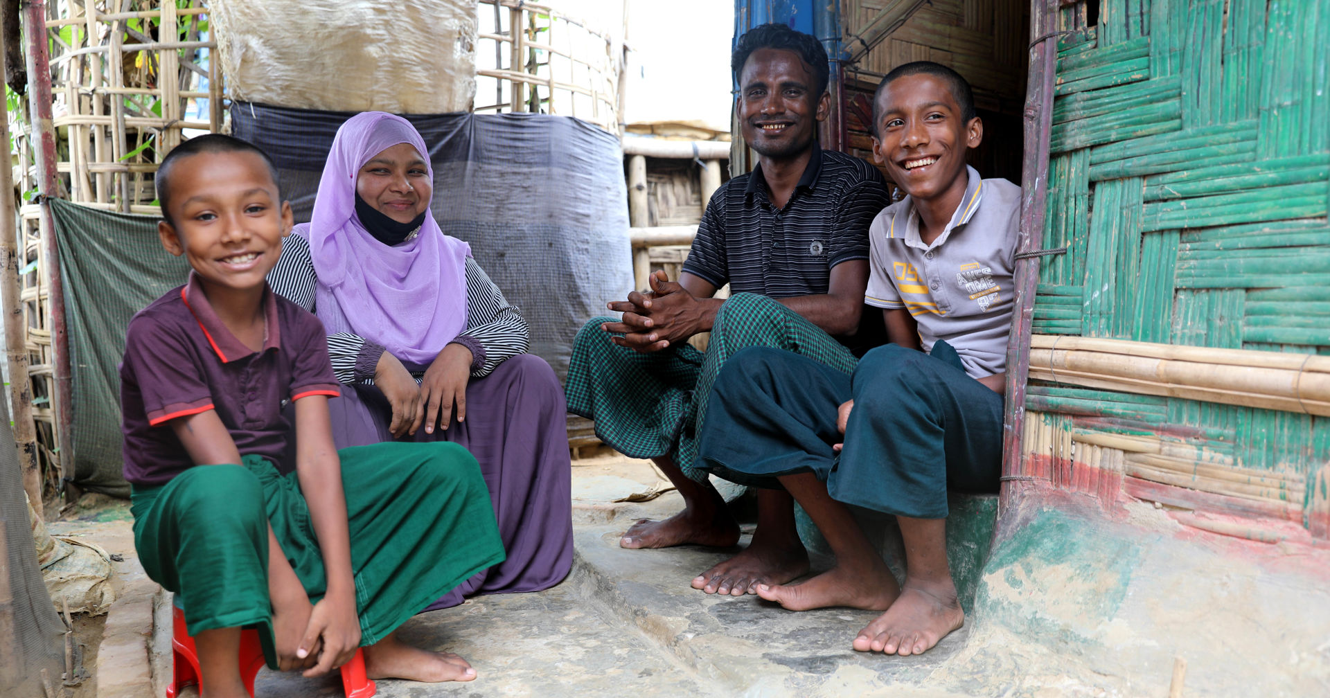 Four people sitting outside and in the doorway in front of dwellings made of bamboo.