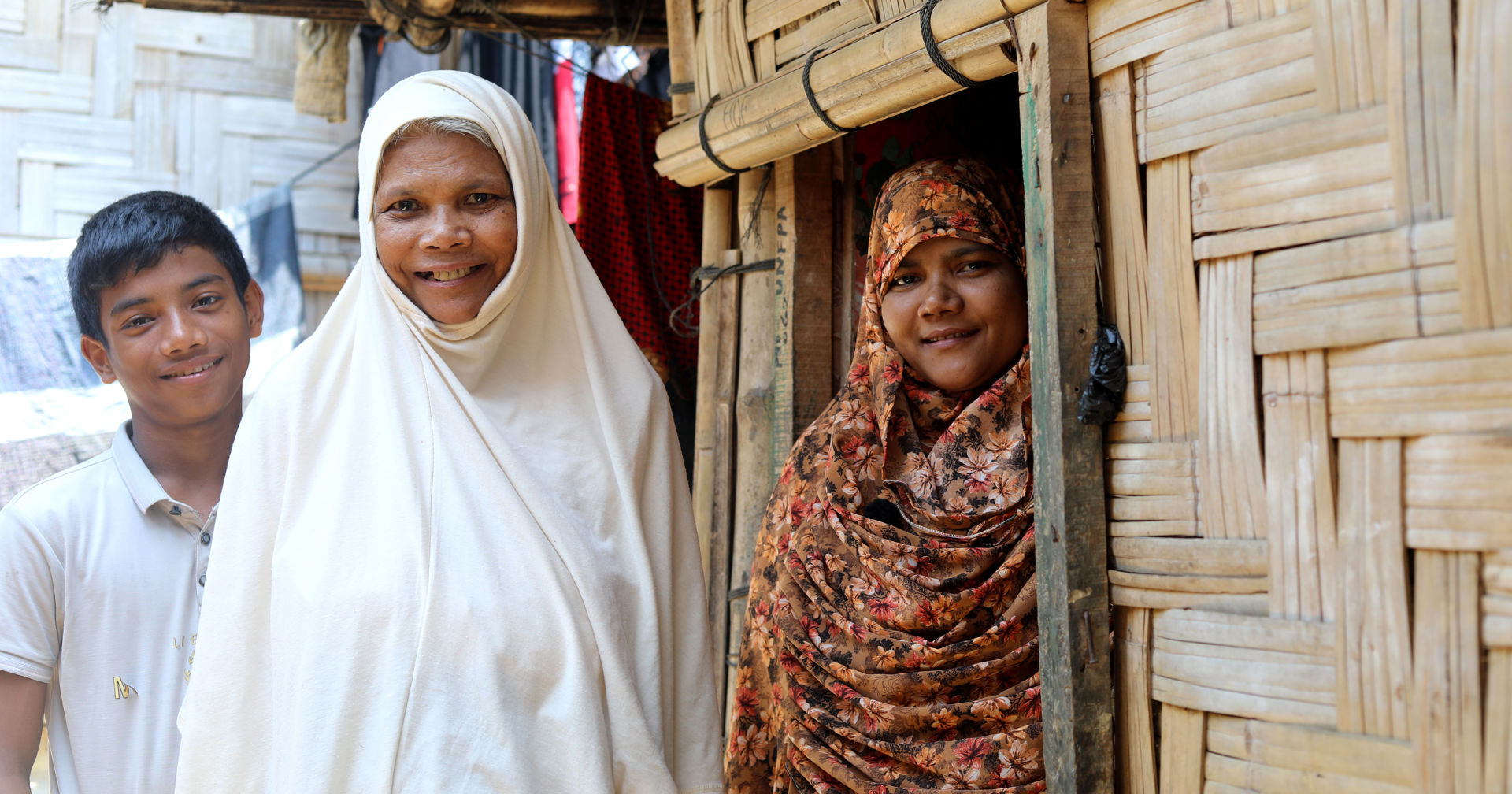 Three people are smiling and looking at the camera from the doorway outside a shelter made of bamboo.