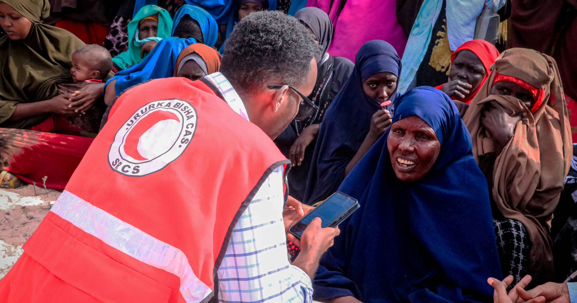 A person wearing a Somali Red Crescent Society vest is leaning towards a person sitting on the ground and asking them questions while holding a mobile phone. 