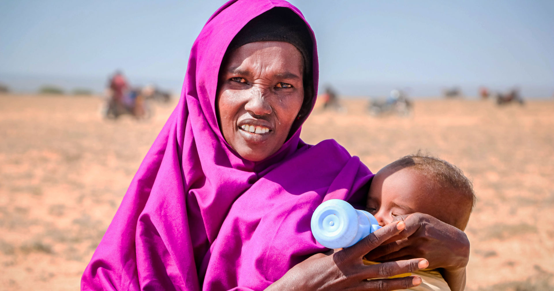 A person wearing a purple scarf is looking at the camera and bottle-feeding a baby in their arms outdoors in the sunshine. 