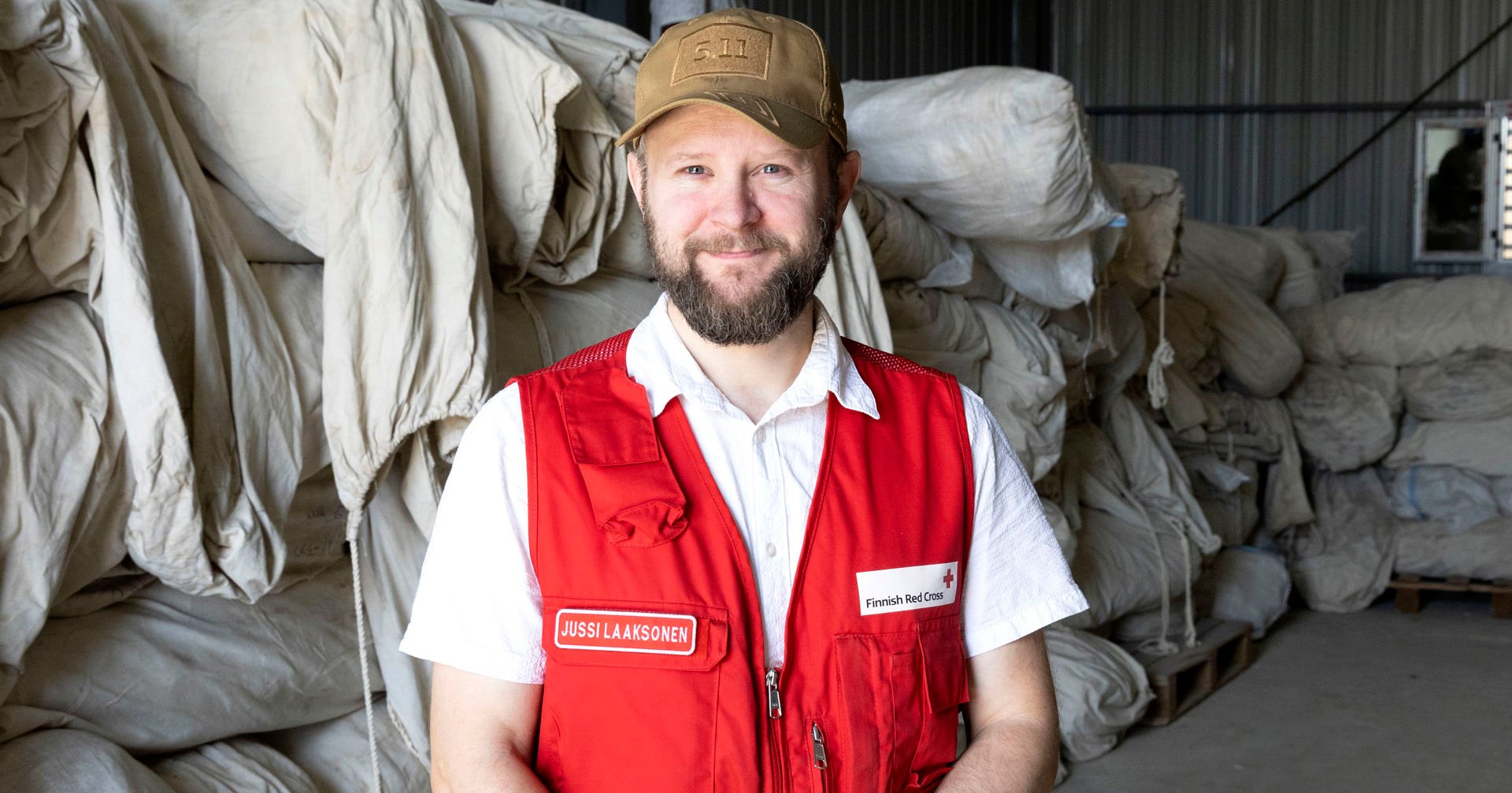 Red Cross logistician Jussi Laaksonen smiles in the storage area. He is wearing a Red Cross work vest, a white shirt, and a brown cap. In the background, there are a large number of tent packages.