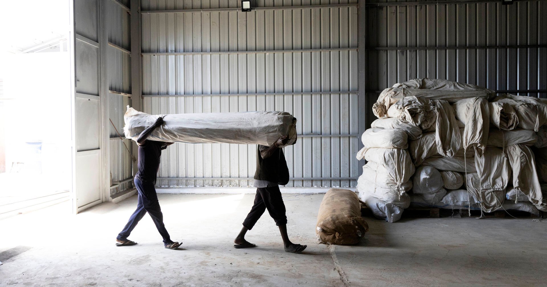 Two people carry a tent covered with a sack into the storage building. In the background, large relief supply bags can be seen.