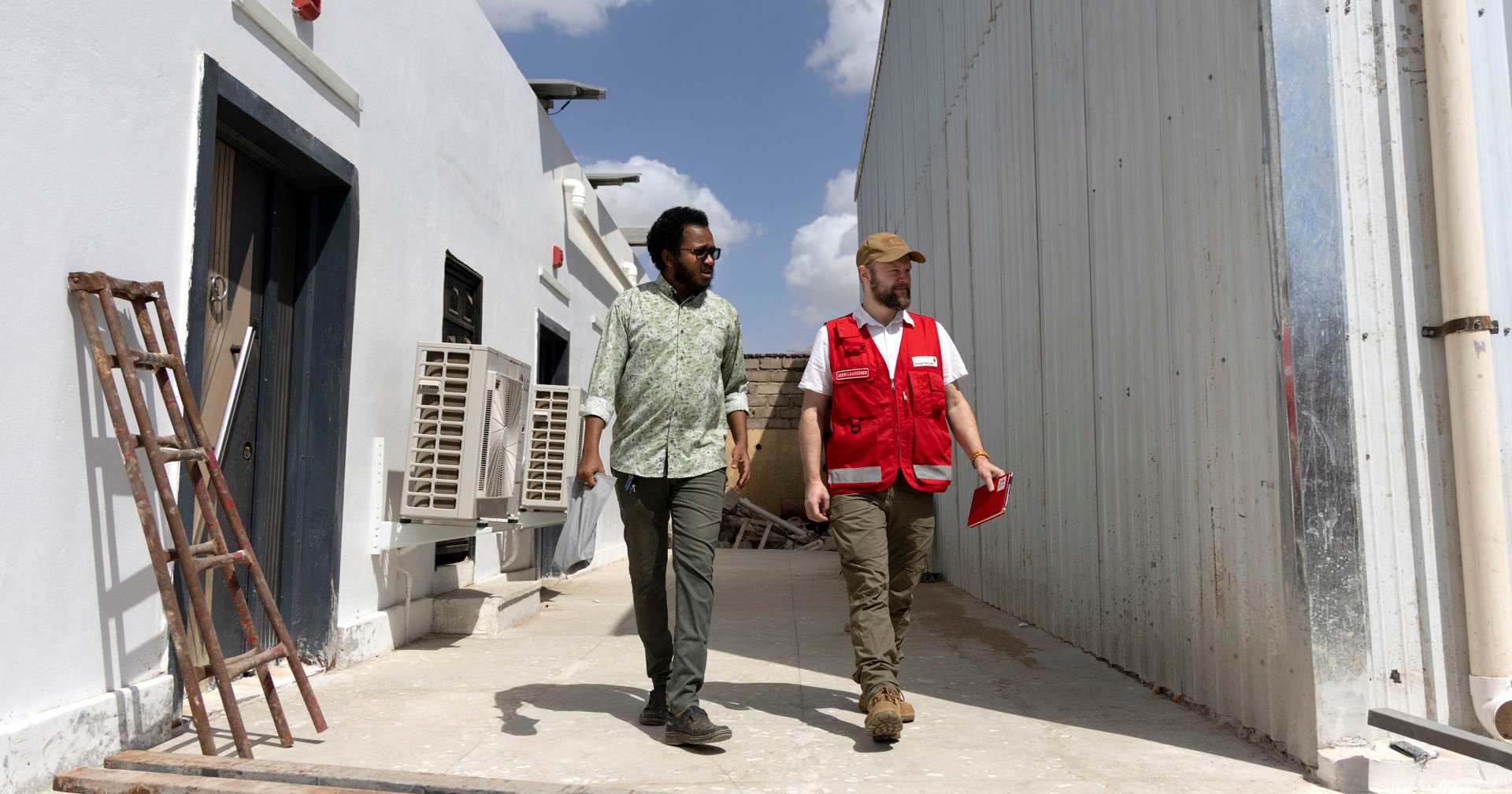 A person wearing a Red Cross vest and another person walk between the storage buildings in the sunny weather.