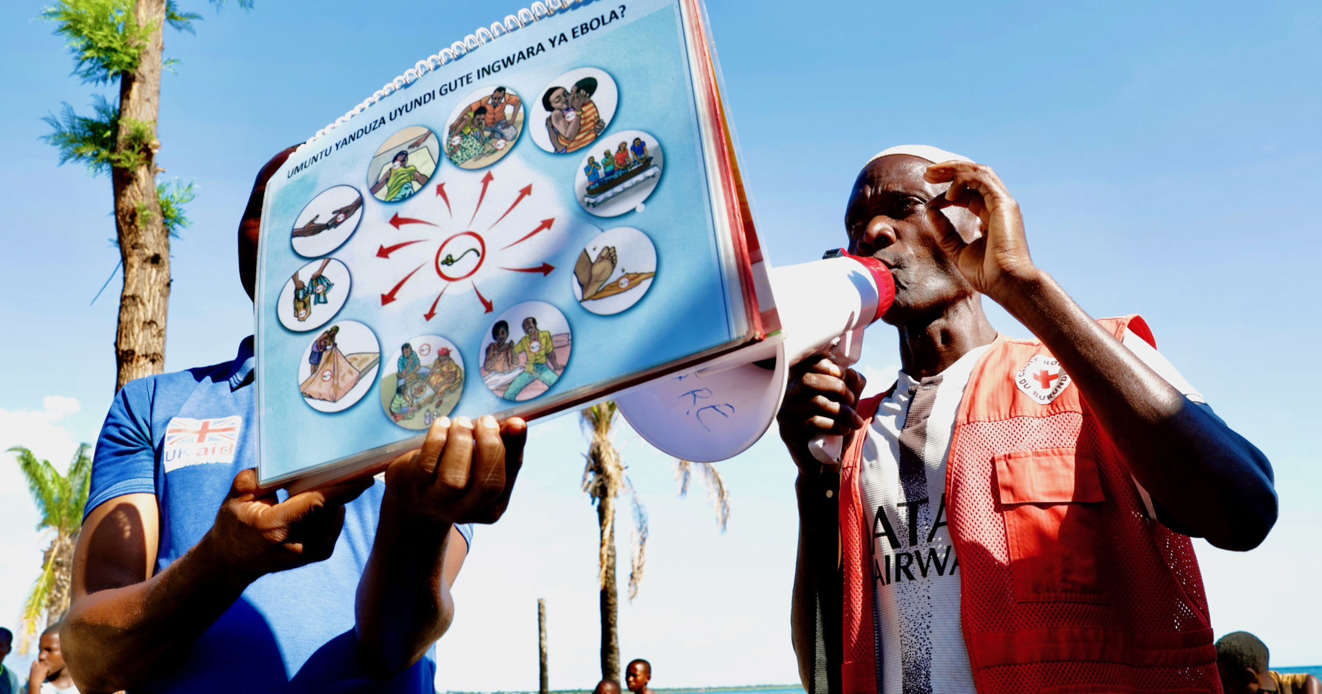 A person wearing a Red Cross vest is speaking into a megaphone, while another person is holding a poster explaining how Ebola is transmitted. 