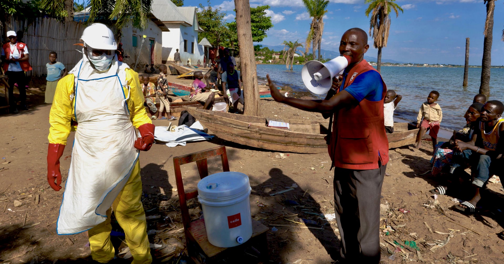 A person wearing a Red Cross vest is speaking into a megaphone on the beach, surrounded by onlookers. Another person is wearing an Ebola protection suit. 