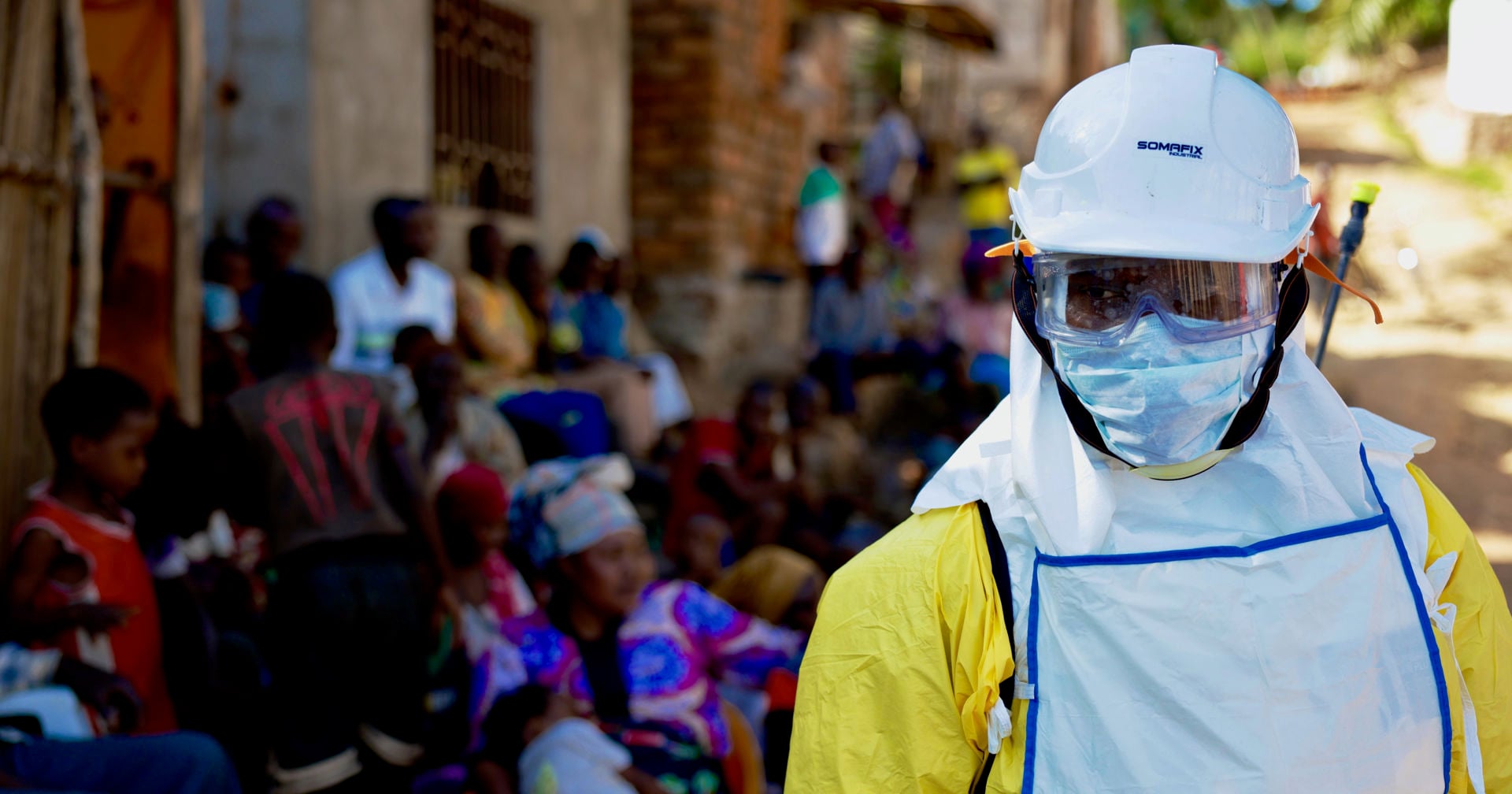 A person wearing an Ebola protective suit looking at the camera, with a group of people sitting in the shade next to a wall in the background.