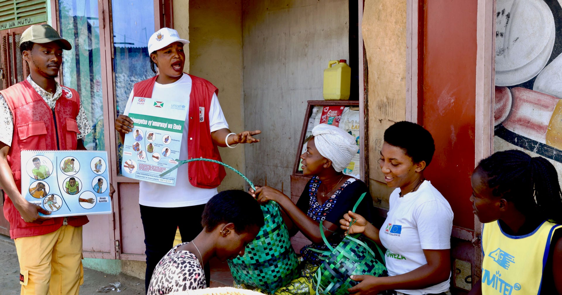 Two people wearing Red Cross vests are using pictures to explain Ebola to people sitting on the street and weaving colourful baskets. 