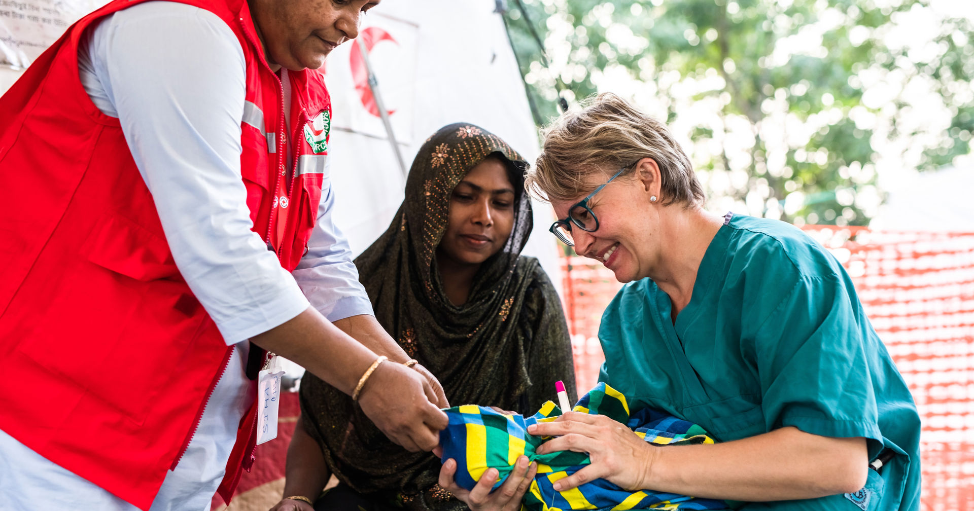 A Red Cross nurse smiles while holding a baby in front of a field hospital tent. Next to them are the baby's mother and a local aid worker.