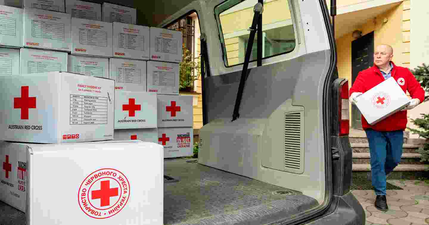 A person in Red Cross uniform carrying a box of Red Cross aid supplies to a van half full of aid supplies.