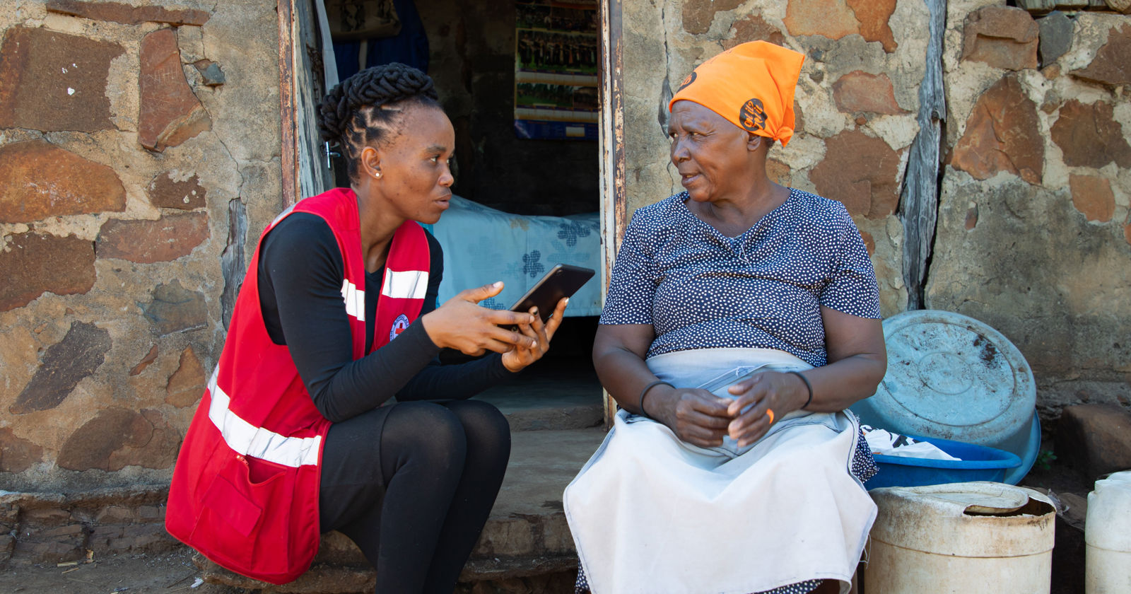 A Red Cross volunteer sits outside a stone house talking with a community member, holding a tablet while they discuss information together.