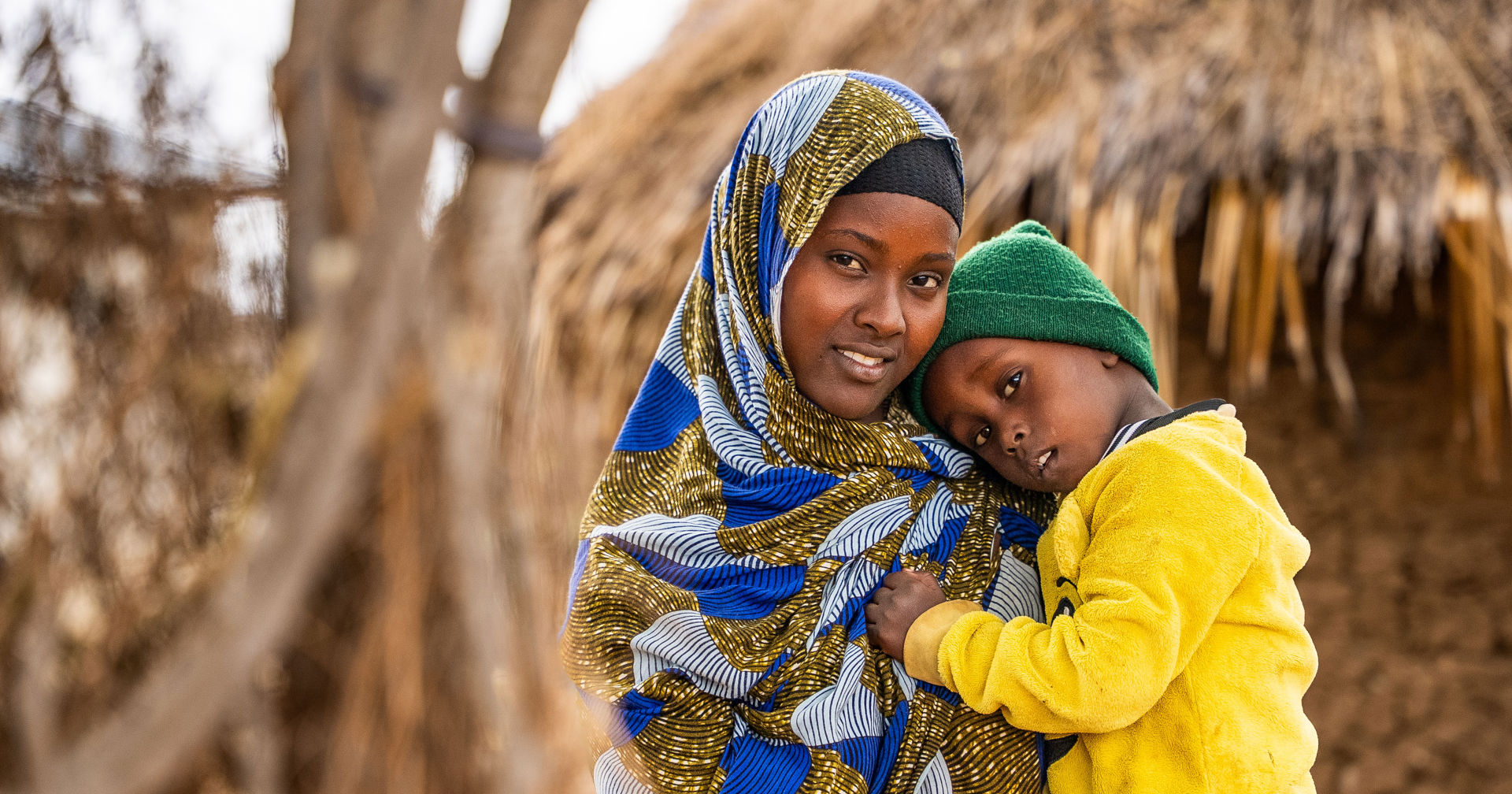 Hawo Yussuf stands in the yard of her home, holding her child in her arms, who is leaning against their mother.