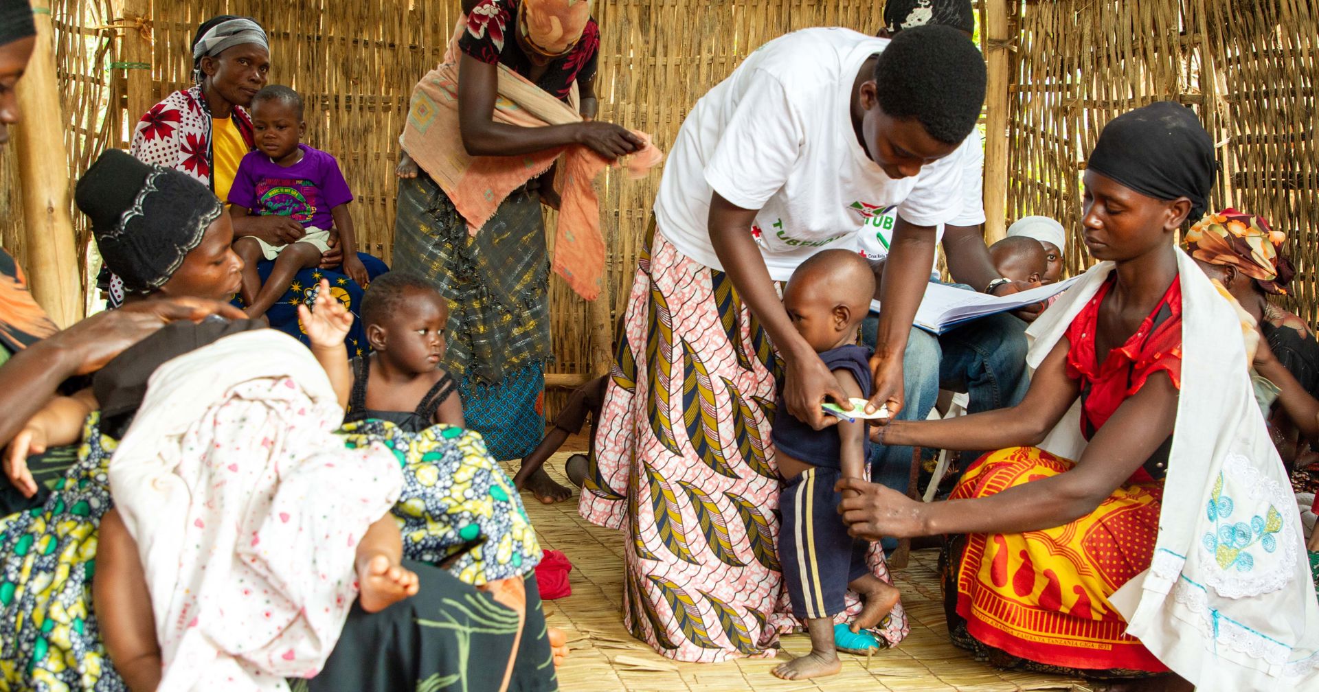 Burundian families at a health post. One person is measuring the circumference of a young child’s upper arm with a measuring tape, with the child’s mother standing nearby.