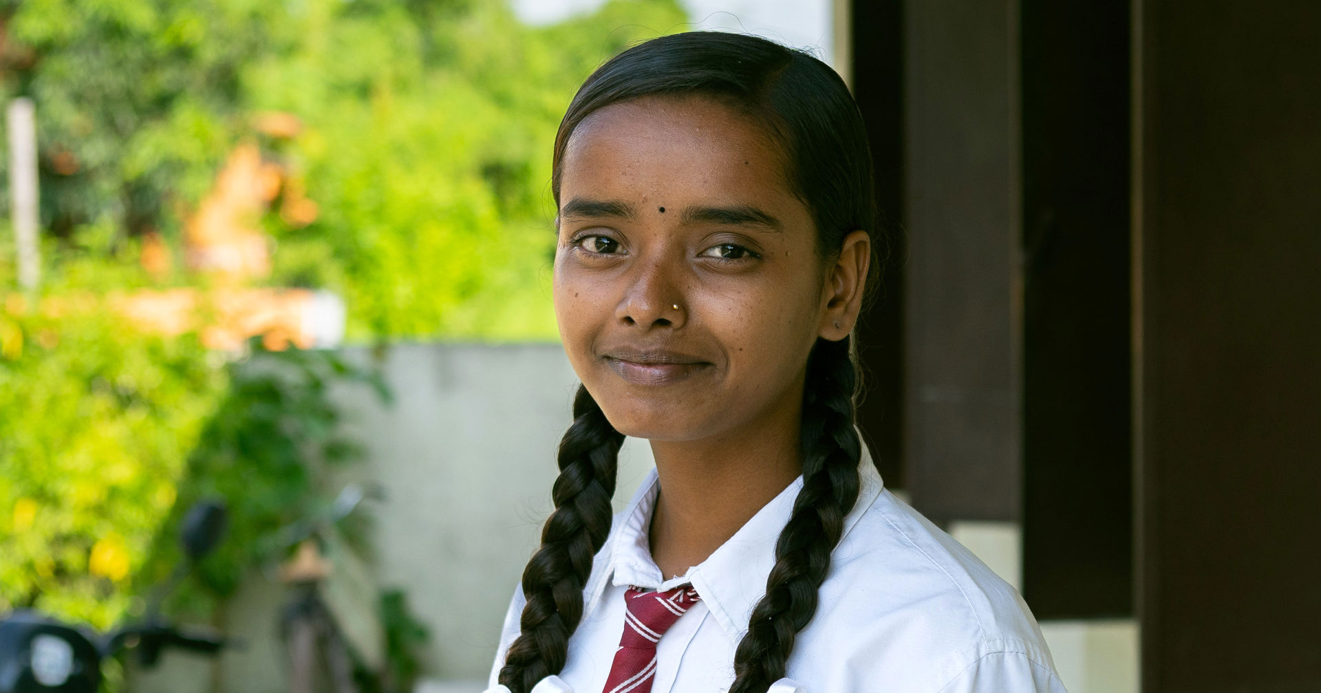 A girl wearing a school uniform, with braided hair, stands outdoors next to a building.