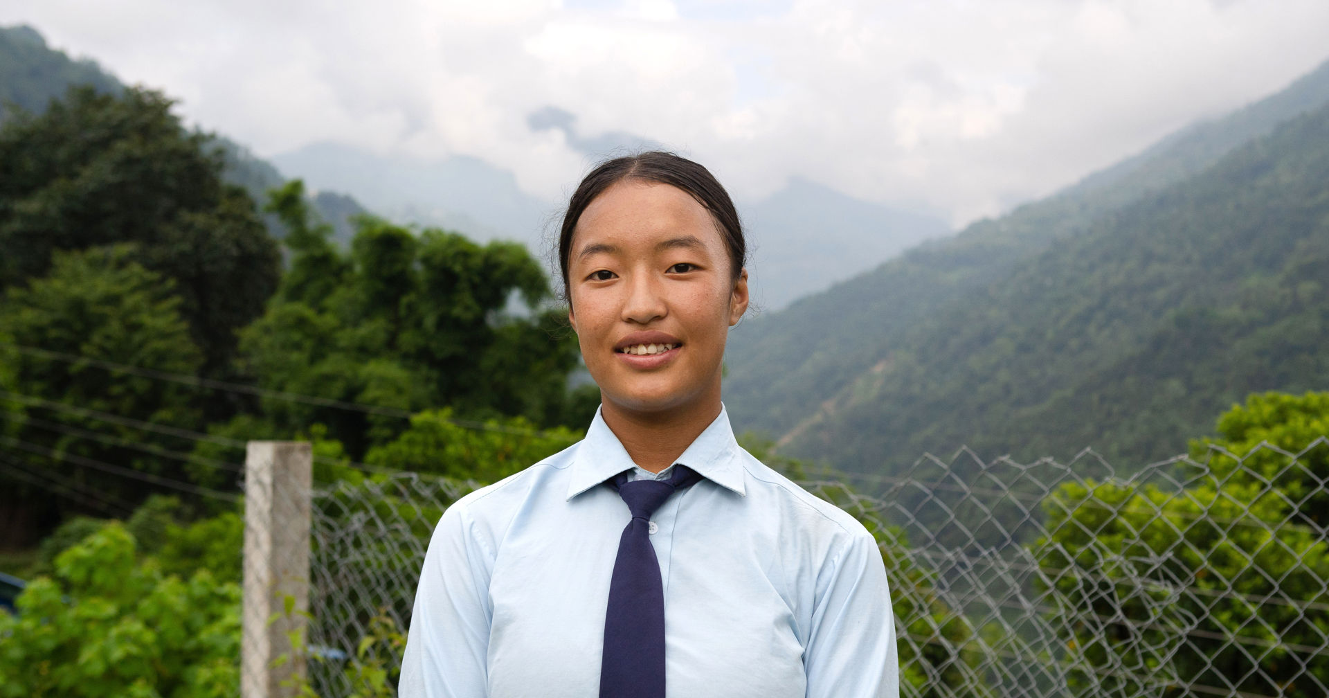 A young person wearing a school uniform stands outdoors in front of a fence. In the background is a green mountainous landscape, trees and clouds.