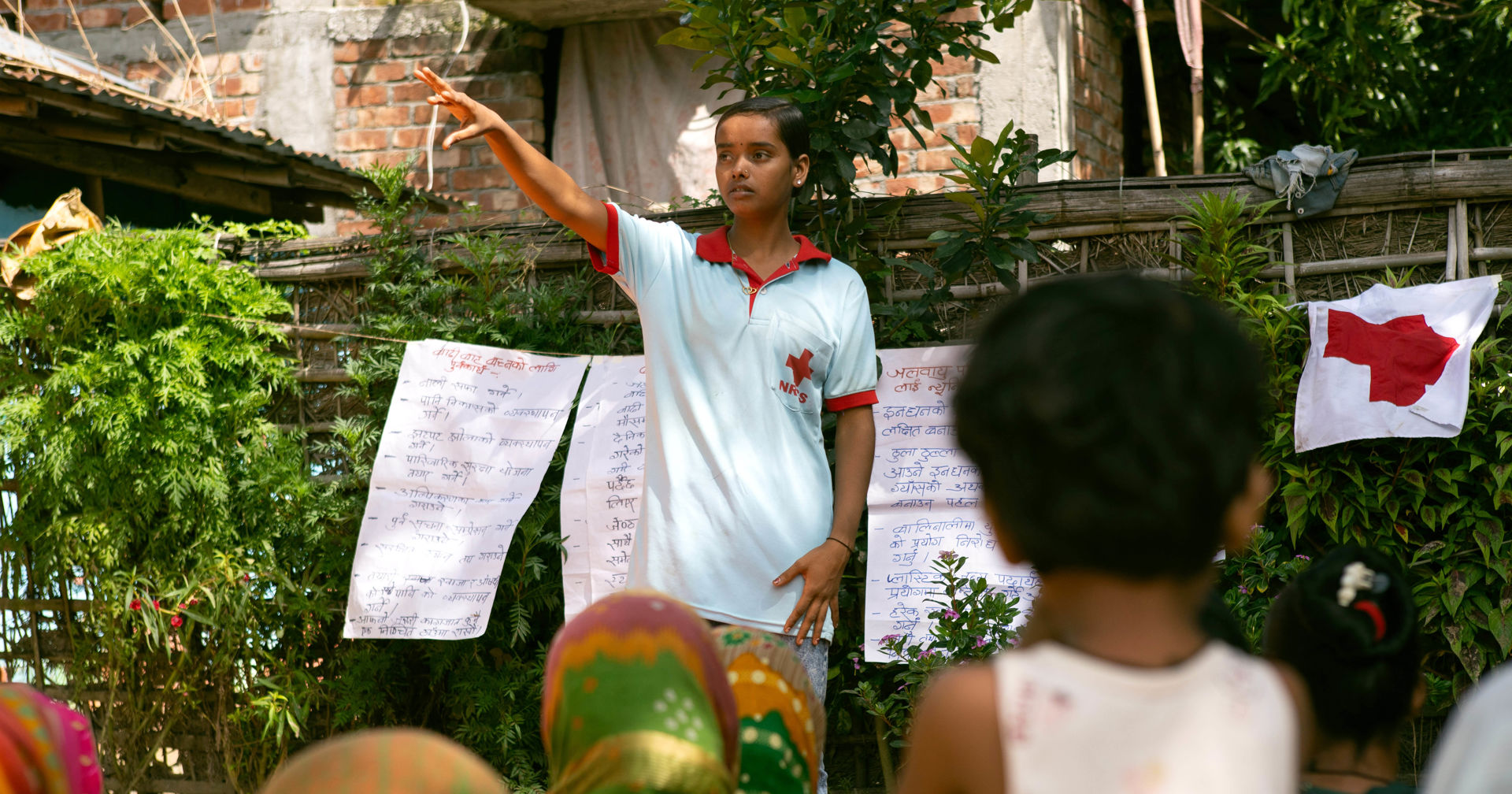 A person stands outdoors wearing a Red Cross T-shirt in front of handwritten posters, pointing forward with one hand while speaking to an audience. A child listens in the foreground, with buildings and vegetation in the background.