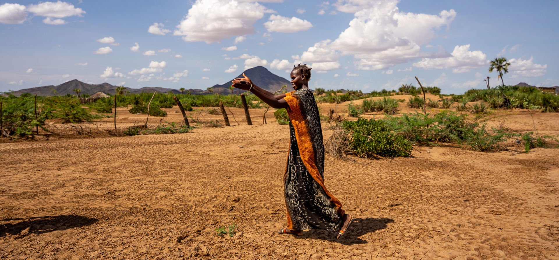 A woman is walking on the orange sand in a village, pointing to a river, which is out of frame in the photo. 
