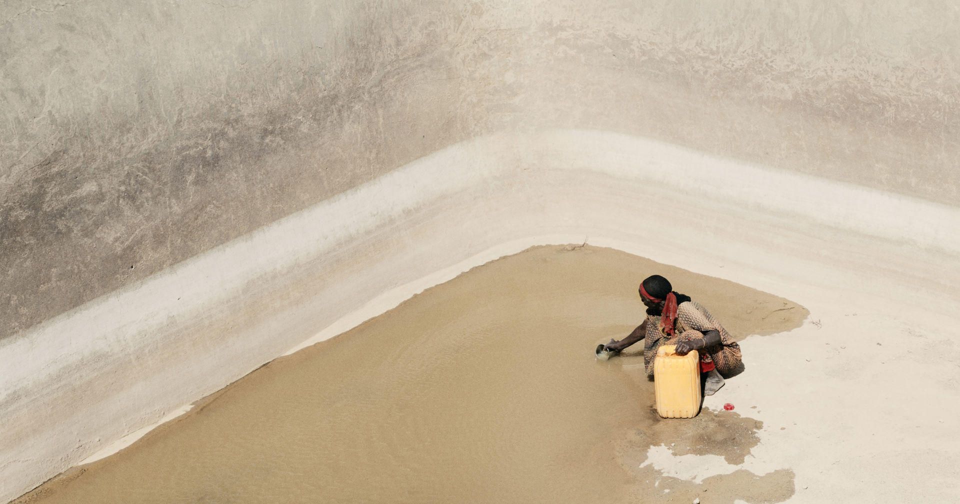 A person fetching water from the bottom of a large water basin. There is only a small puddle left at the bottom of the basin.