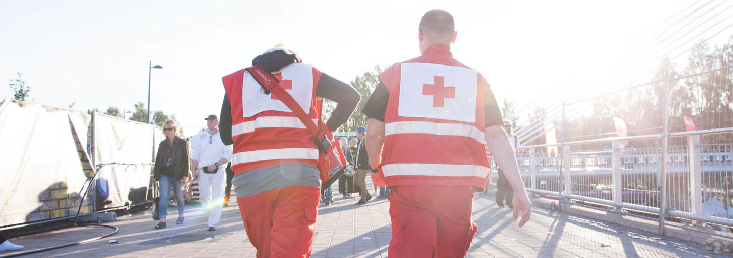 On first aid duty at an outdoor event, a man and a woman wearing Red Cross uniforms walk towards the sunset.