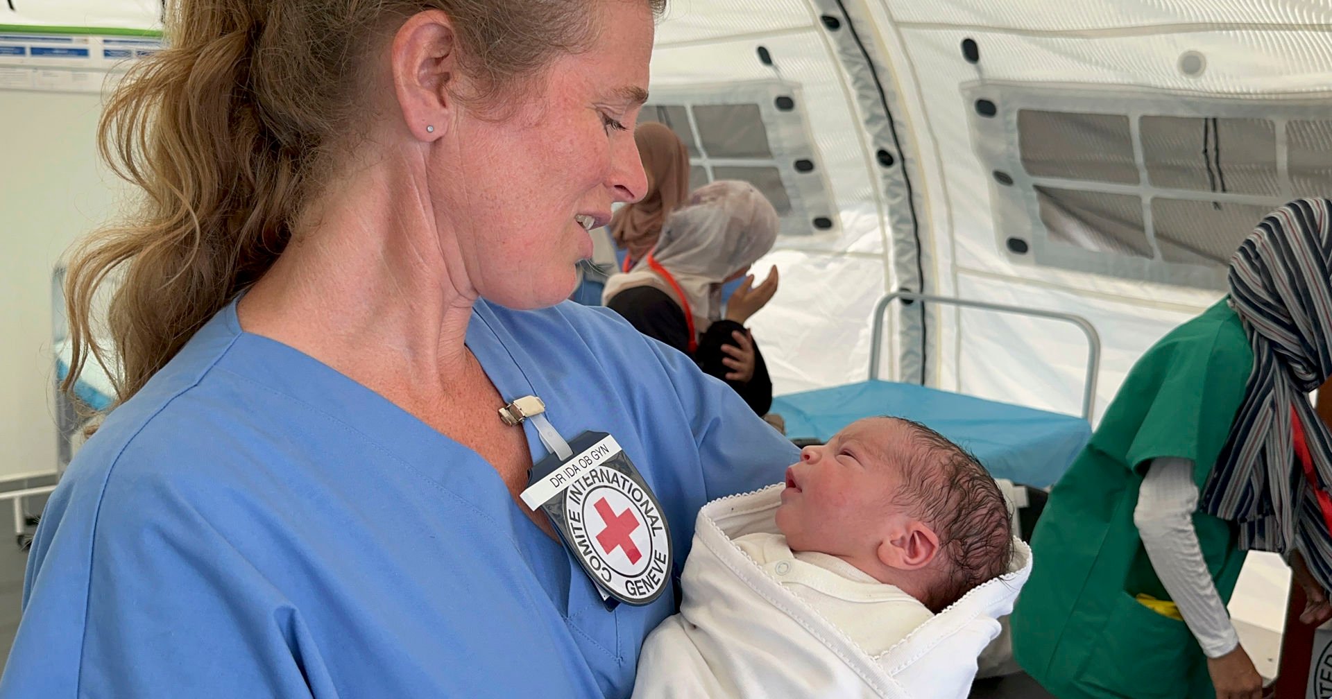 A healthcare professional wearing blue scrubs is holding a newborn baby. The person has an International Red Cross badge and an ID card on the chest. In the background, the interior of a medical tent is visible, along with an examination table and other individuals in protective clothing.
